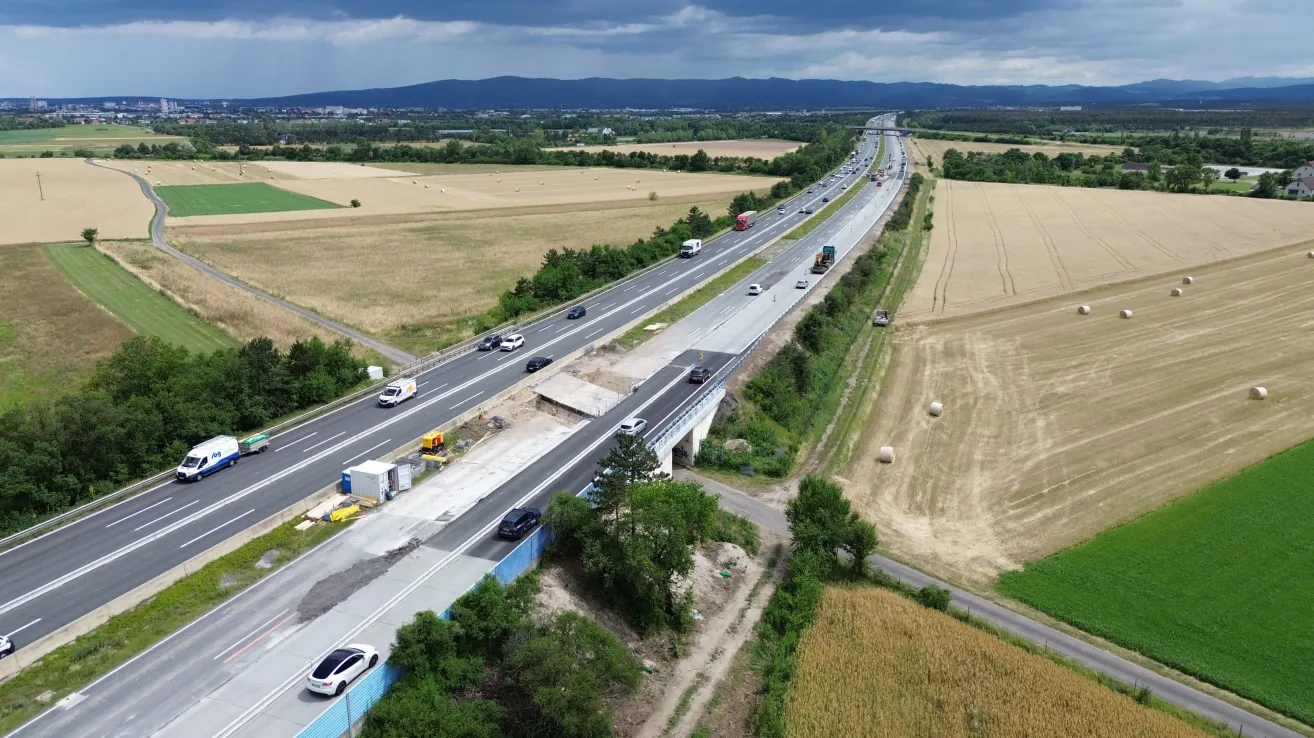 An aerial view of a busy highway with vehicles driving on both sides, surrounded by green fields and mountains in the distance.