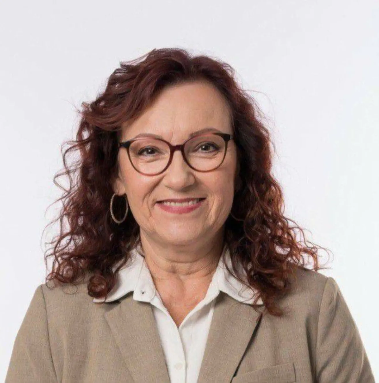 A woman with curly brown hair wearing glasses and a beige blazer smiles for a photo against a white background.