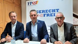 Three men in suits sit at a table in front of a banner with German text. The man in the middle smiles.