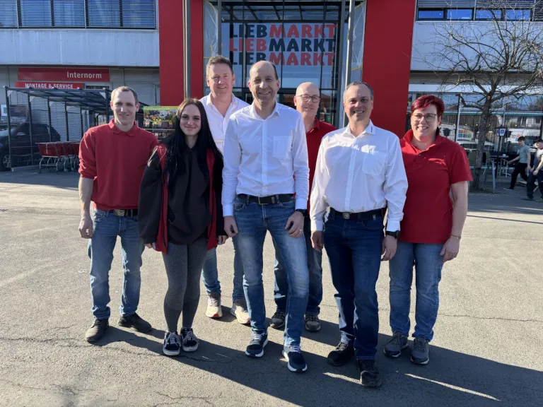 A group of six people are standing in front of a Liebmarkt store, all wearing red shirts and smiling for a photo.