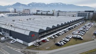 An aerial view of a large industrial building with a parking lot filled with cars. Mountains are visible in the distance.