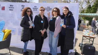 Four women, each holding a book, stand in front of a white banner with German text, smiling and posing for a photo. They are dressed in winter clothing, and behind them is a building with several windows.
