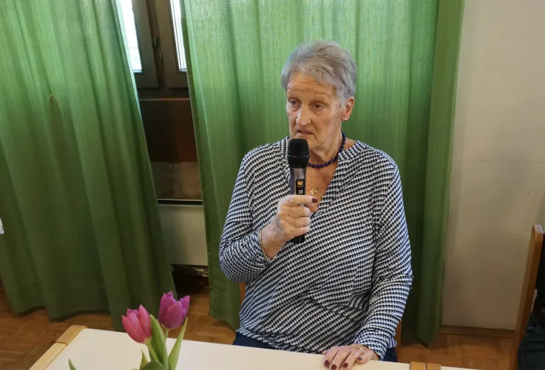 An elderly woman speaks into a microphone at a table with a flower arrangement, against a backdrop of green curtains.