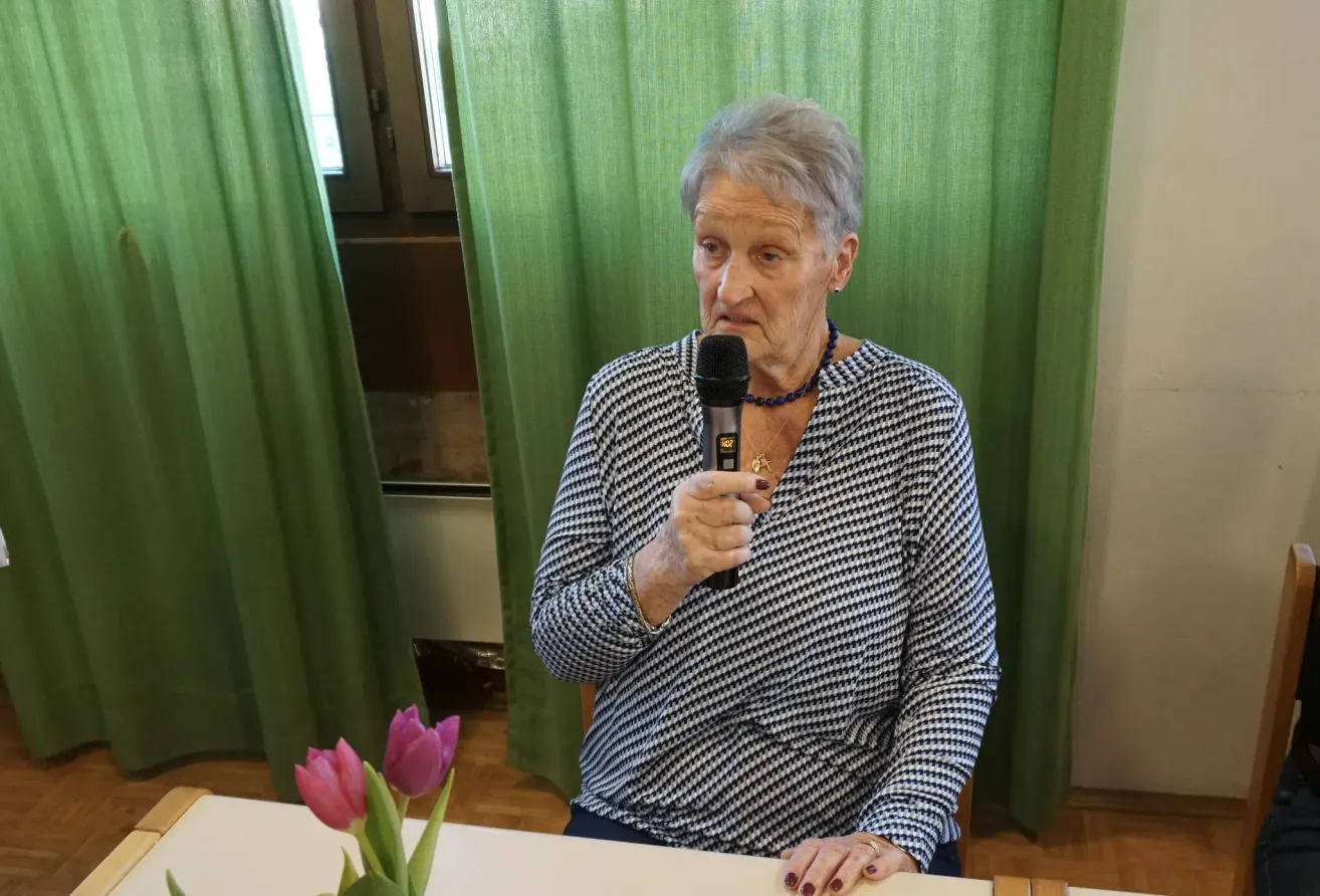 An elderly woman speaks into a microphone at a table with a flower arrangement, against a backdrop of green curtains.
