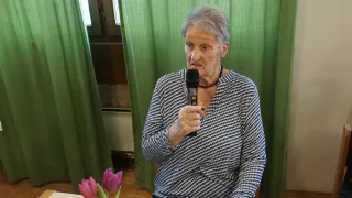 An elderly woman speaks into a microphone at a table with a flower arrangement, against a backdrop of green curtains.