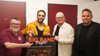 Four men stand behind a poster for the Big Band Project. The man in the center holds the poster with both hands. Behind them, two red doors are visible.