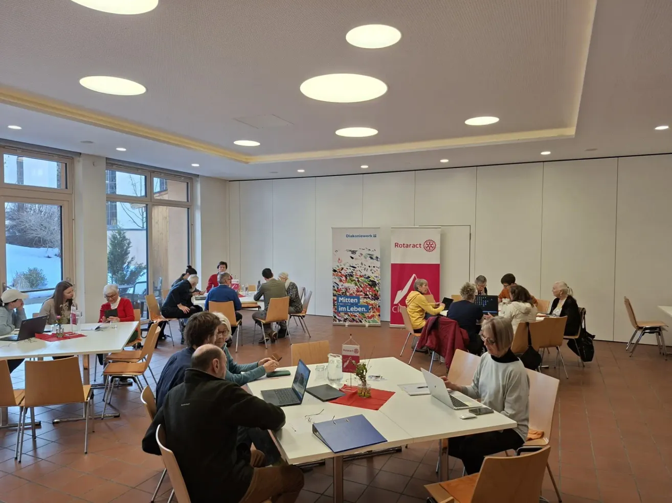 A group of people are sitting around tables in a room. Some of them are using laptops, while others are reading papers. The room has white walls and round ceiling lights. There are two banners on the wall. The floor is covered with tiles.
