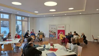 A group of people are sitting around tables in a room. Some of them are using laptops, while others are reading papers. The room has white walls and round ceiling lights. There are two banners on the wall. The floor is covered with tiles.