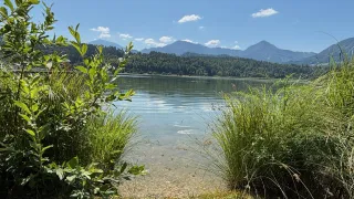 A serene lake with clear water and a grassy shoreline is seen under a blue sky with scattered clouds, surrounded by lush greenery and distant mountains.