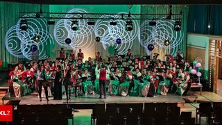 A large orchestra on stage with green and red attire performs under a green backdrop with spiral designs and balloons.