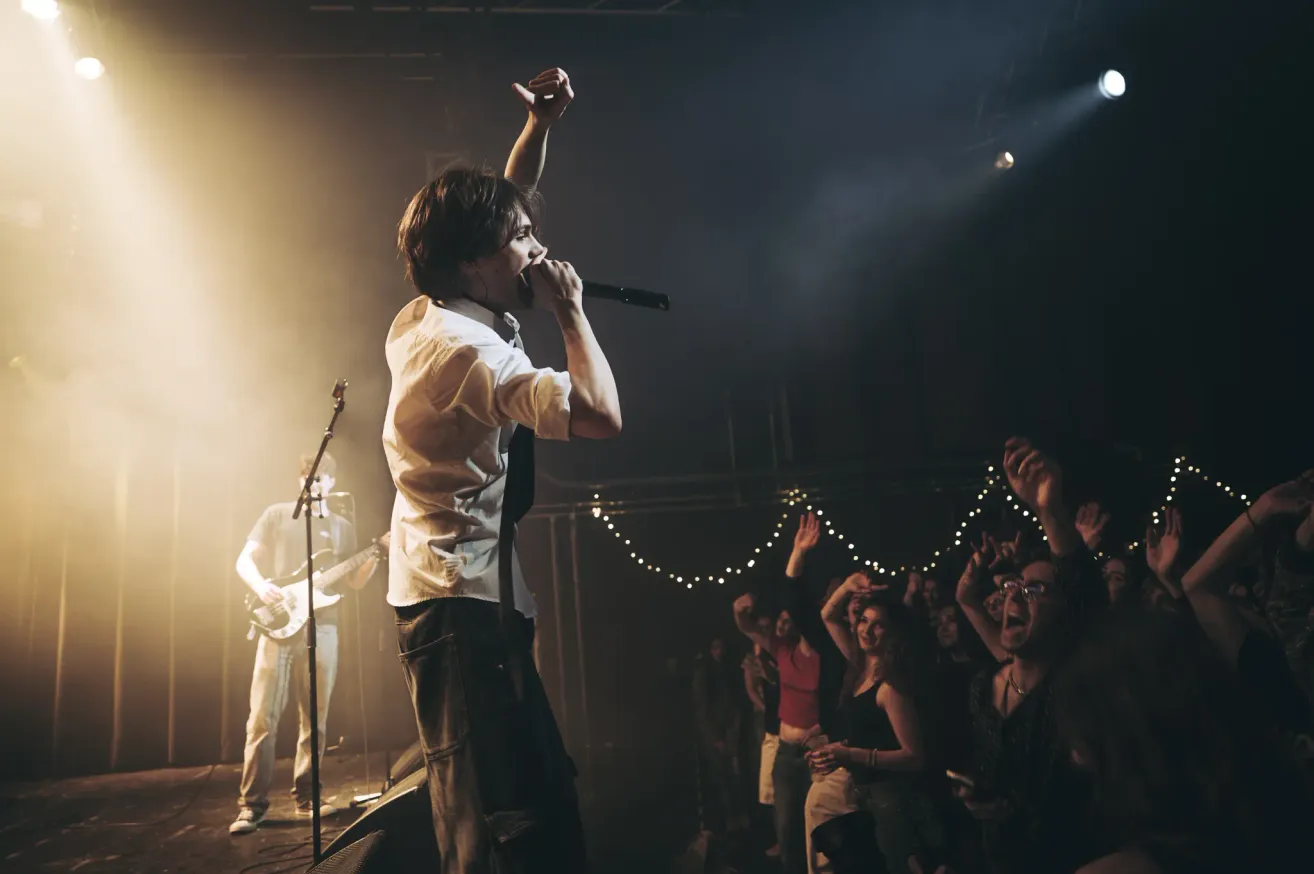 A rock band performs on stage with a singer singing into a microphone while a guitarist plays in the background. The crowd is cheering and raising their hands.