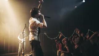 A rock band performs on stage with a singer singing into a microphone while a guitarist plays in the background. The crowd is cheering and raising their hands.