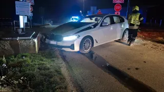 A white car is parked on the side of the road at night with a damaged front. A person in a reflective vest and helmet stands near the car.