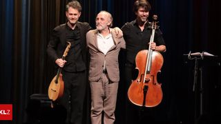 Three musicians on stage, two holding string instruments, one holding a mandolin, smiling and posing for a photo.