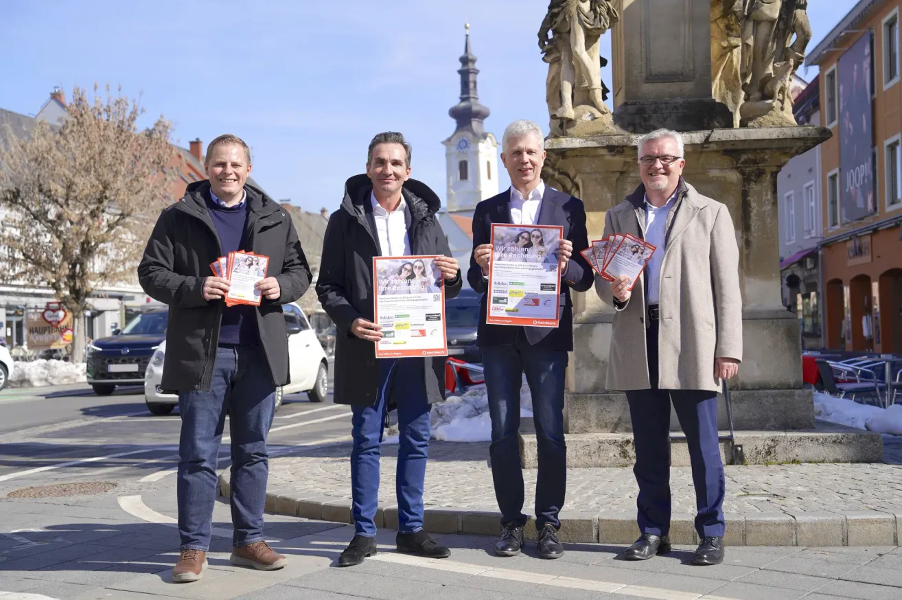 Four men in winter coats stand in front of a monument, each holding brochures. They are smiling and appear to be engaged in a conversation. Behind them is a church with a steeple, and parked cars are visible in the background.