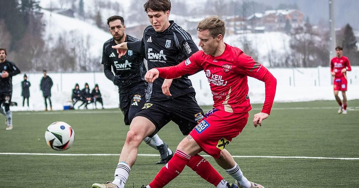 Three soccer players in action on a snowy field. One player in red is tackling another in black. In the background, two more players sit on a bench.