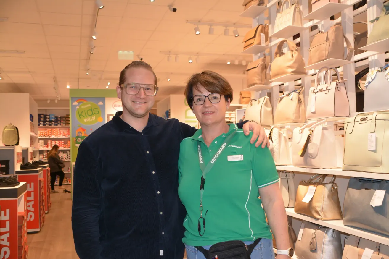 A man and a woman in a store with a shelf full of handbags. The man is smiling while the woman has a fanny pack.