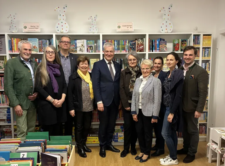 Eine Gruppe von Menschen, darunter Erwachsene und Kinder, steht in einer Bibliothek. Sie alle lächeln und posieren für ein Foto. Hinter ihnen stehen Bücherregale voller Bücher. Einige der Bücher haben ein Etikett, das Kinderbücher sagt.