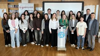 A group of young adults, possibly students, stands in a room. They are in front of a whiteboard with an advertisement. They are all wearing formal attire. A podium with a logo is in front of them.