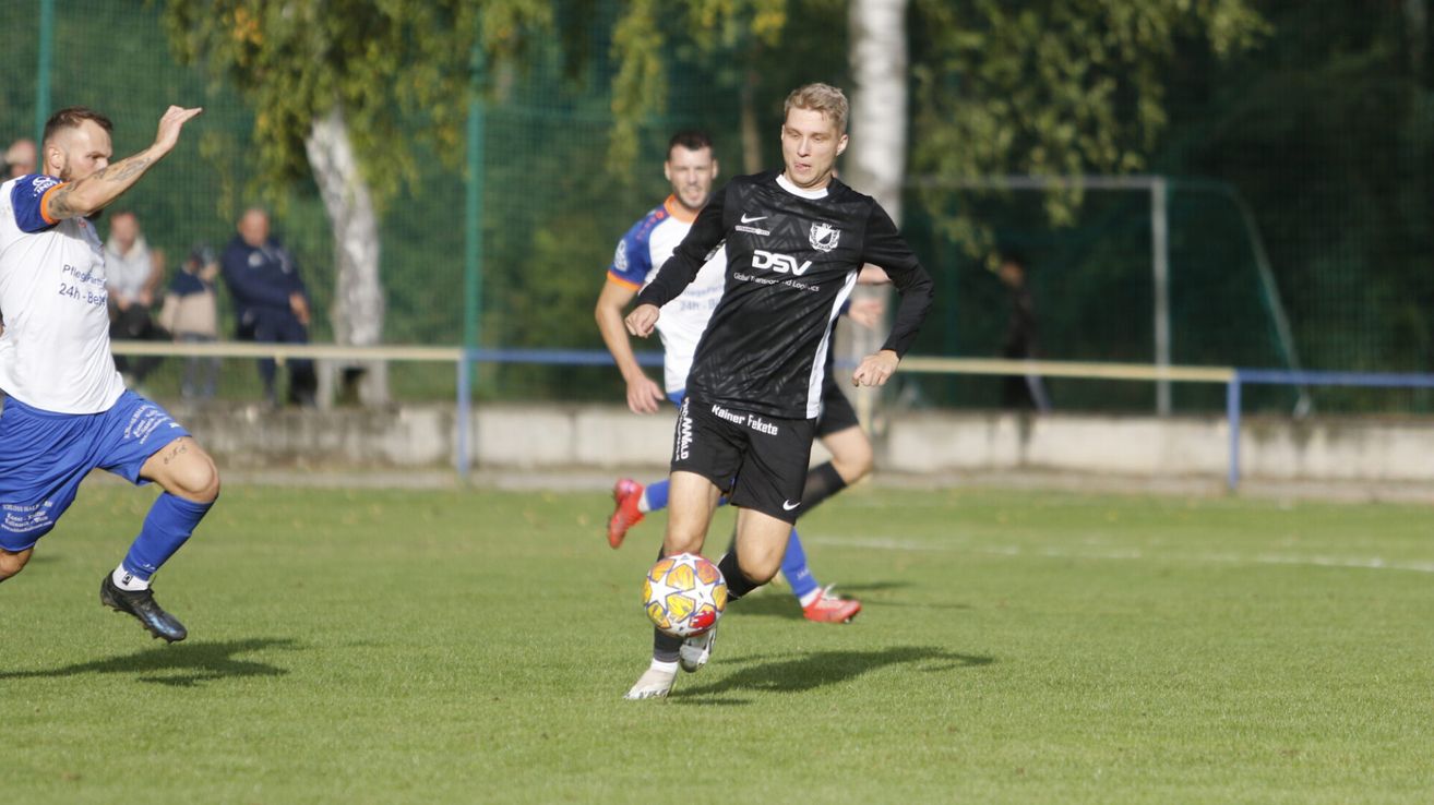 A soccer player in black uniform with DSV on the jersey kicks the ball on a grassy field with another player behind him.