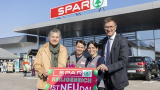 A group of people stand in front of a Spar store, holding a sign with flowers and balloons. A man and two women smile and pose for a photo.