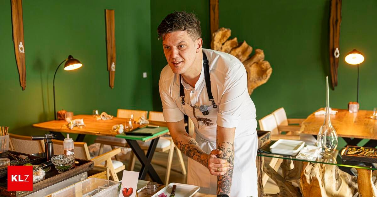 A tattooed chef in a white apron stands in a restaurant with green walls, holding a piece of paper with a heart on it.