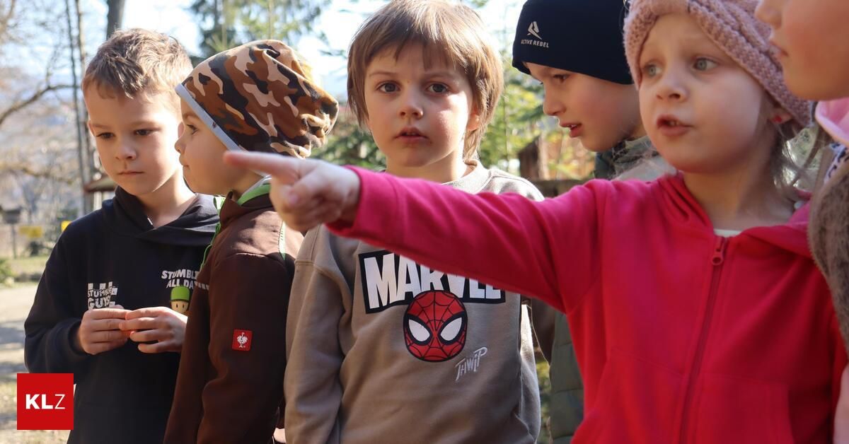 A group of children stand in a line; one child points at something. The child in the center wears a Marvel sweatshirt, while others wear beanie hats.