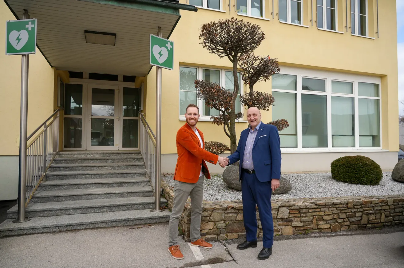 Two men in business suits stand in front of a building, one shaking hands with the other. The building has a green sign with a heart and lightning bolt.