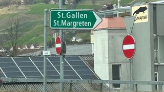 A street sign indicates directions to St. Gallen and St. Margreten. It is mounted on a pole next to a building with a solar panel on its roof.