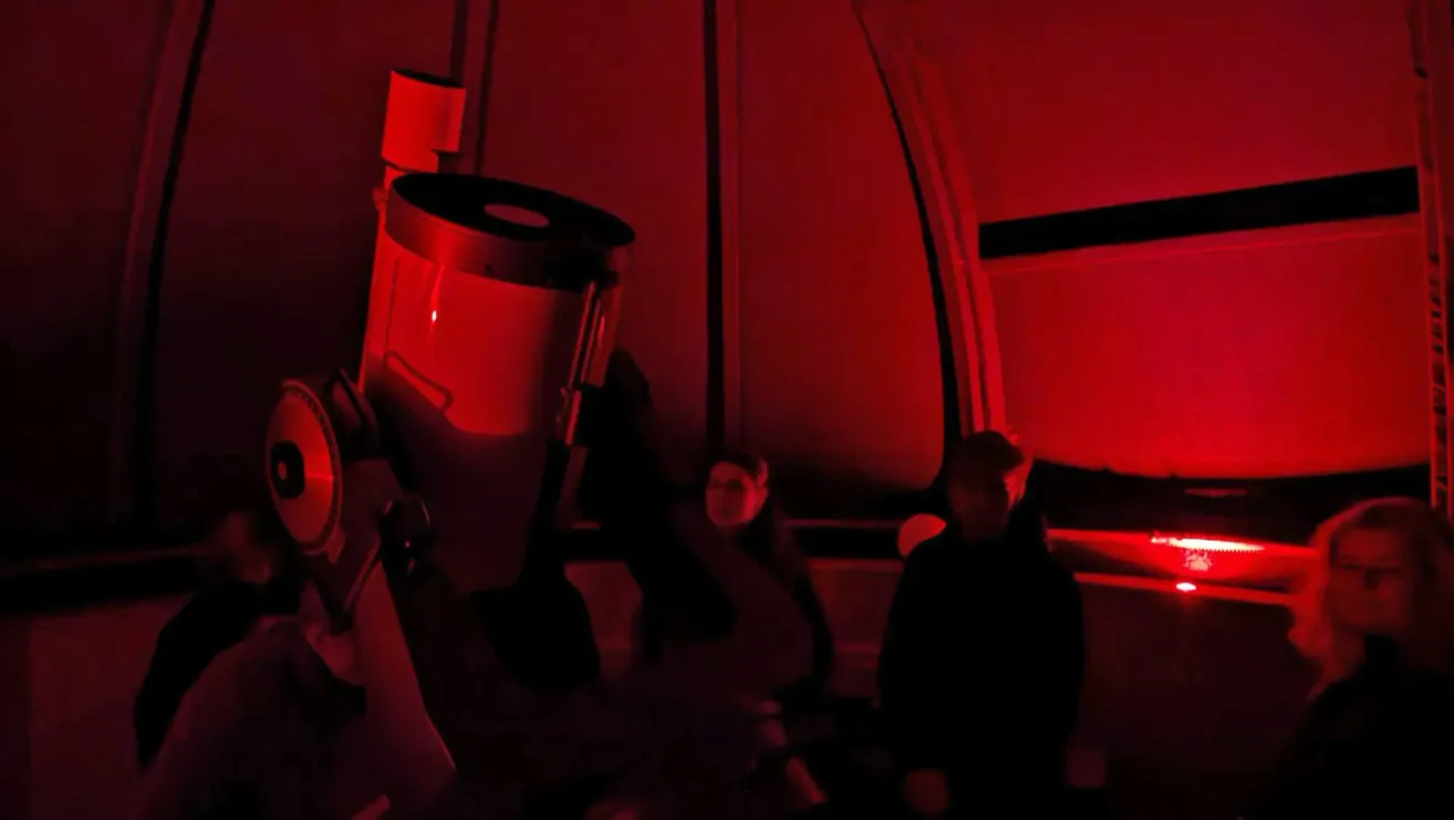 In a dimly lit room, a large telescope is set up with red lighting. Two individuals are seated nearby, observing through the telescope.