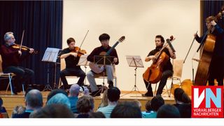 Four musicians are performing on stage, with three playing string instruments and one seated audience member taking a photo.