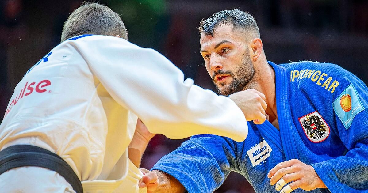 Zwei Männer in Kampfsportuniformen, einer in Blau und der andere in Weiß, befinden sich in einer engen Grappling-Position. Der Mann in Blau hat einen konzentrierten Ausdruck und einen Bart.