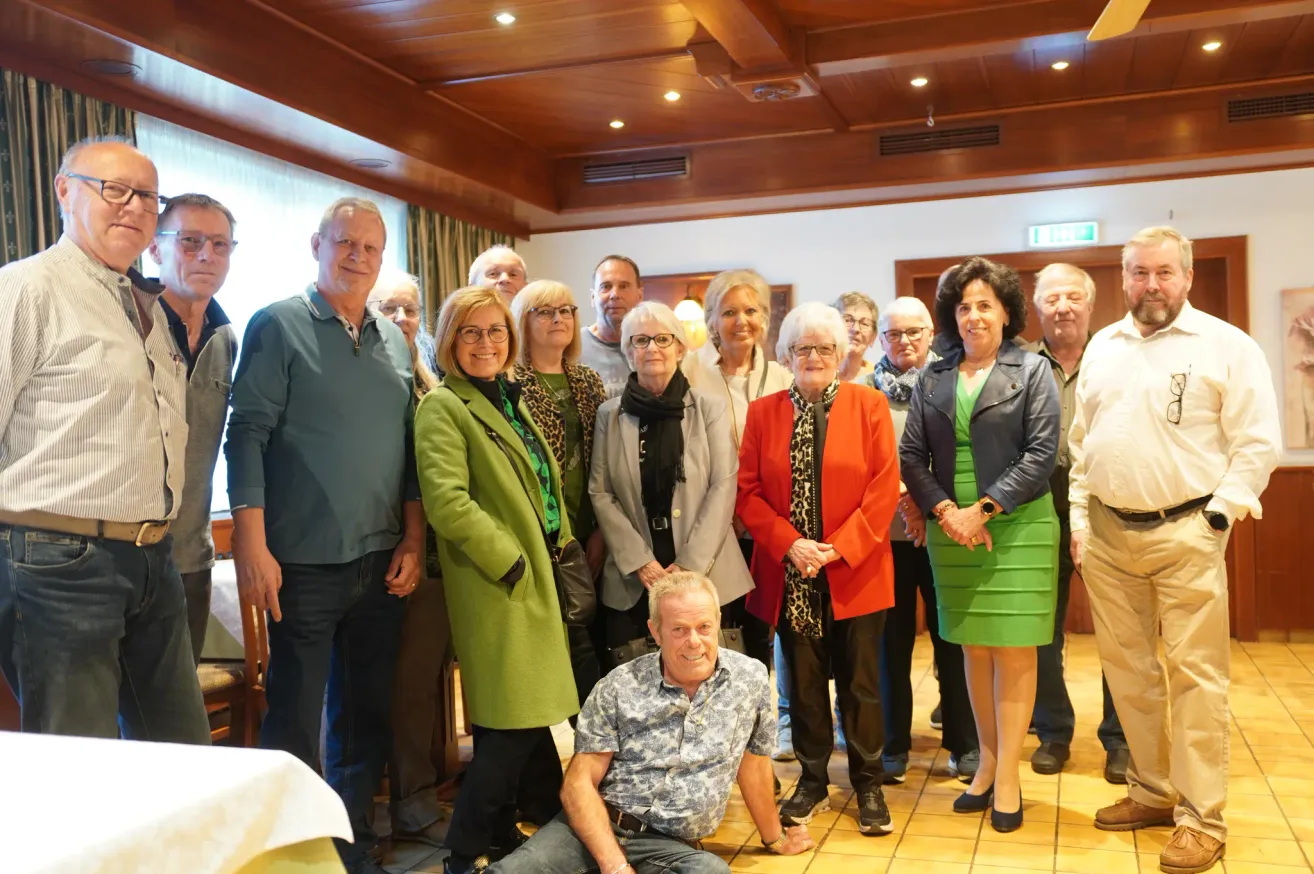 A group of people, including men and women, are standing and sitting in a room. Some are wearing glasses and smiling, while others are looking at the camera. They are likely posing for a group photo. The room has a tiled floor and a wooden ceiling.
