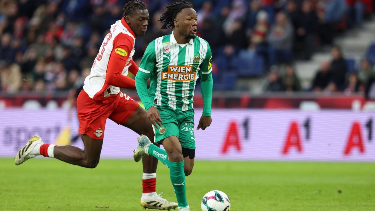 Two soccer players on a field, one wearing a green and white uniform with 'WIEN ENERGIE' and the other in red and white. They are chasing a soccer ball.