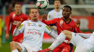 Two soccer players in white jerseys are in a tussle for the ball. The player in white wears a logo for Dormero Hotels. Another player in red has a Puma logo on his jersey.
