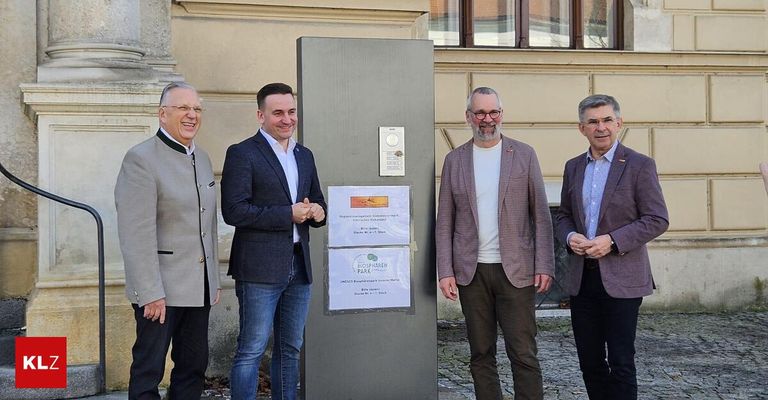 Four men are standing next to a gray post with signs on it, smiling and talking. They are dressed formally and are in front of a building with large windows.