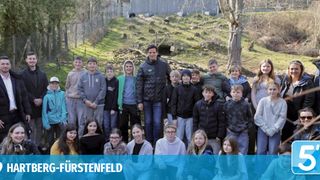 A group of young people and an adult man pose for a photo in a forest. They stand in front of a fence and a house in the background.