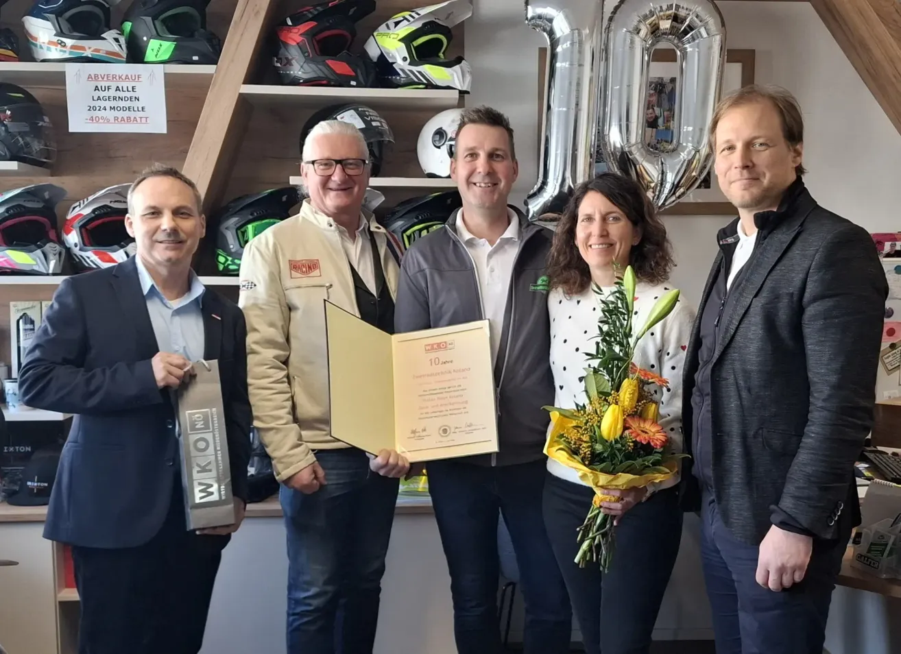 Fünf Erwachsene posieren für ein Foto, um einen Meilenstein zu feiern, mit einem Zertifikat, Blumen und einem Nummer-10-Ballon. Der Mann links hält einen Pokal, und sie stehen vor Regalen mit Helmen und einem weißen Schild.