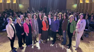 A group of women in formal attire, some wearing glasses, are standing on a stage with flowers in front of them. They seem to be posing for a photograph. Behind them, there is a large crowd of people sitting on chairs.
