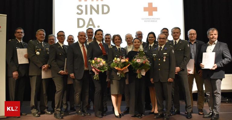 A group of people in formal attire pose on a stage with flowers, behind them a screen displays 'Wir sind da.' and the Red Cross logo.