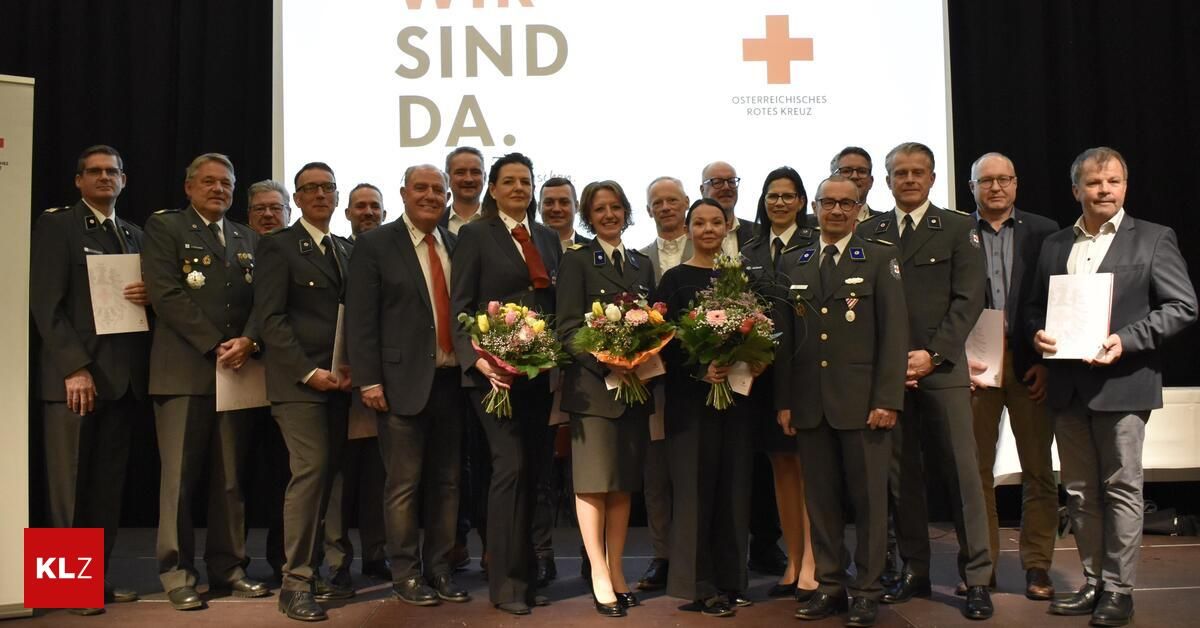 A group of people in formal attire pose on a stage with flowers, behind them a screen displays 'Wir sind da.' and the Red Cross logo.