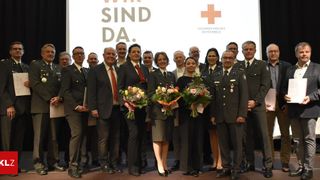 A group of people in formal attire pose on a stage with flowers, behind them a screen displays 'Wir sind da.' and the Red Cross logo.