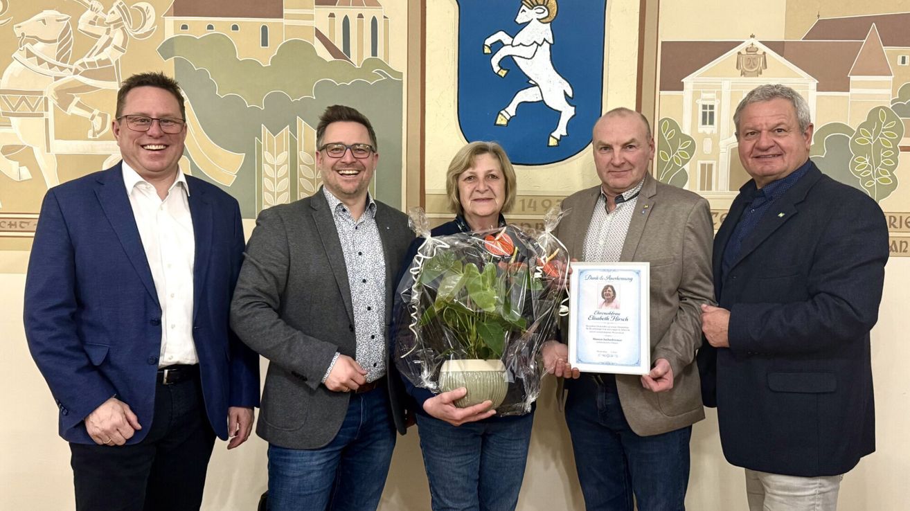 Drei Menschen stehen eng beieinander, lächeln, einer hält ein gerahmtes Zertifikat und eine Topfpflanze. Hinter ihnen ist eine Wand mit einem blau-weißen Emblem.