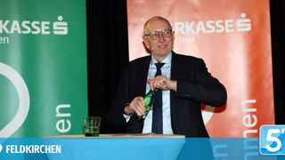 An older man in a suit is standing in front of a table, holding a green bottle and pouring its contents into a glass. Behind him is a red backdrop with text.