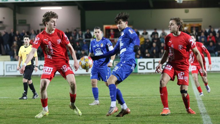 Vier Fußballspieler konkurrieren auf einem Feld, Zuschauer im Hintergrund. Zwei Spieler in blauen Trikots fordern einen Spieler in einem roten Trikot heraus, der einen Fußball hält.