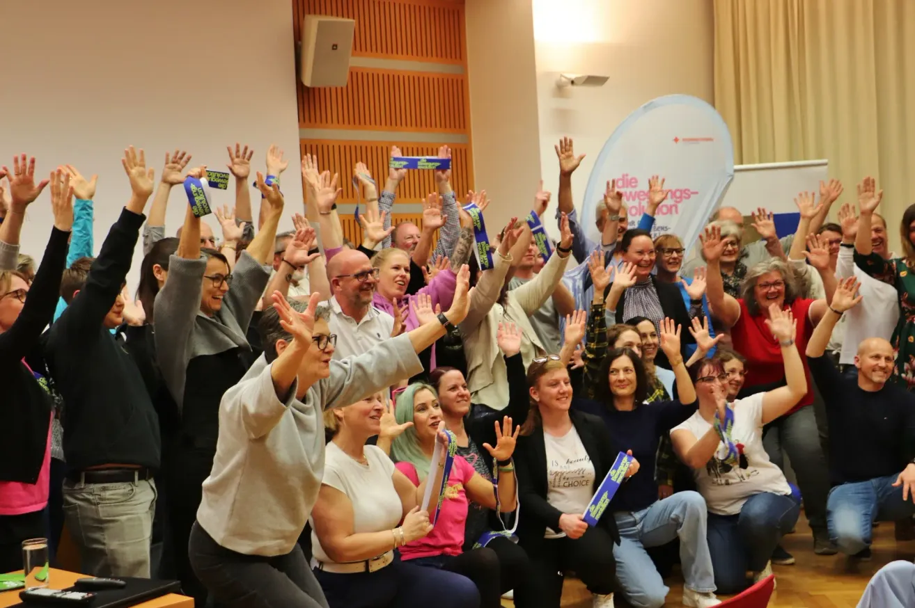A group of people in a hall, raising their hands and smiling. Some hold ribbons. A banner with text is visible on the wall.
