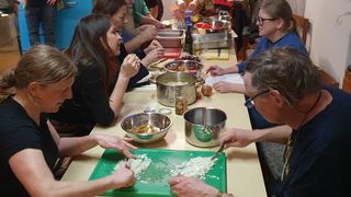 A group of people are seated around a table, engaging in a cooking activity. They are chopping onions on a green cutting board and there are various pots and ingredients on the table.