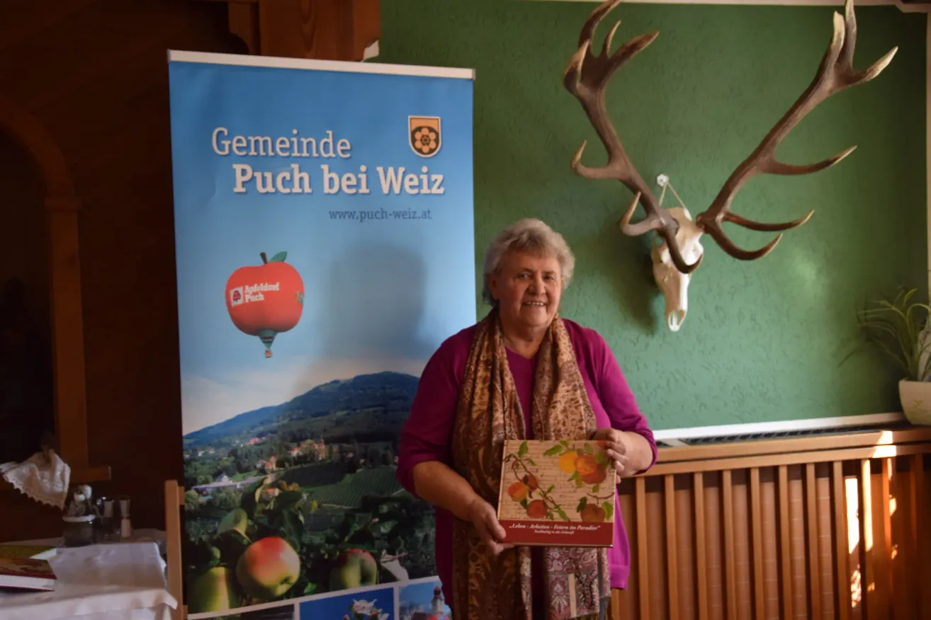 An older woman stands in front of a green wall, holding a box of apples. Behind her is a poster advertising 'Puch bei Weiz', featuring a hot air balloon and a deer head.