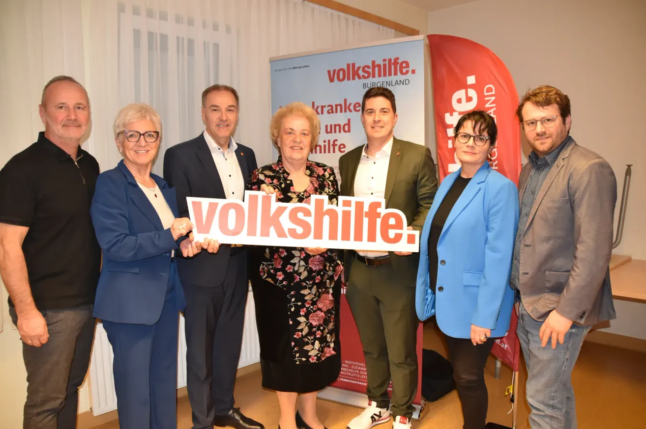 A group of people in formal attire are posing for a photo. They are holding a sign that reads 'volkshilfe.' Behind them, a banner displays the text 'kranke und hilfe.' The setting appears to be an indoor event.