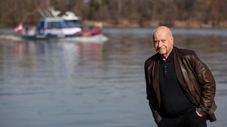 An elderly man in a brown leather jacket stands near the water, smiling with a boat in the background.